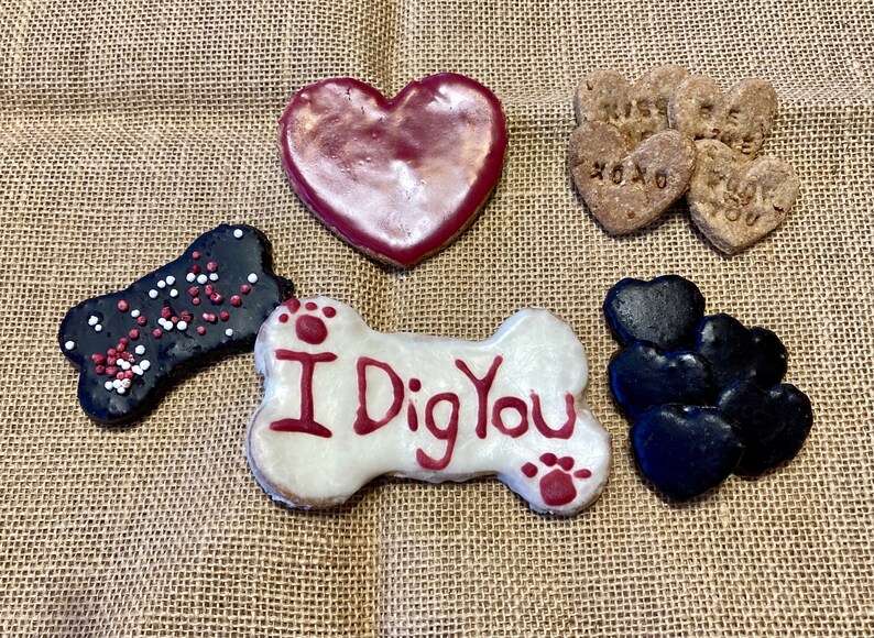 May include: Assortment of dog treats in the shape of hearts and bones. The treats are decorated with red, white and black icing and sprinkles. One bone-shaped treat has the words "I Dig You" written on it.