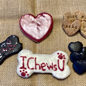 May include: Assortment of dog treats in the shape of hearts and bones. The treats are decorated with red, white and black icing and sprinkles. One bone-shaped treat has the words "I Chews U" written on it.