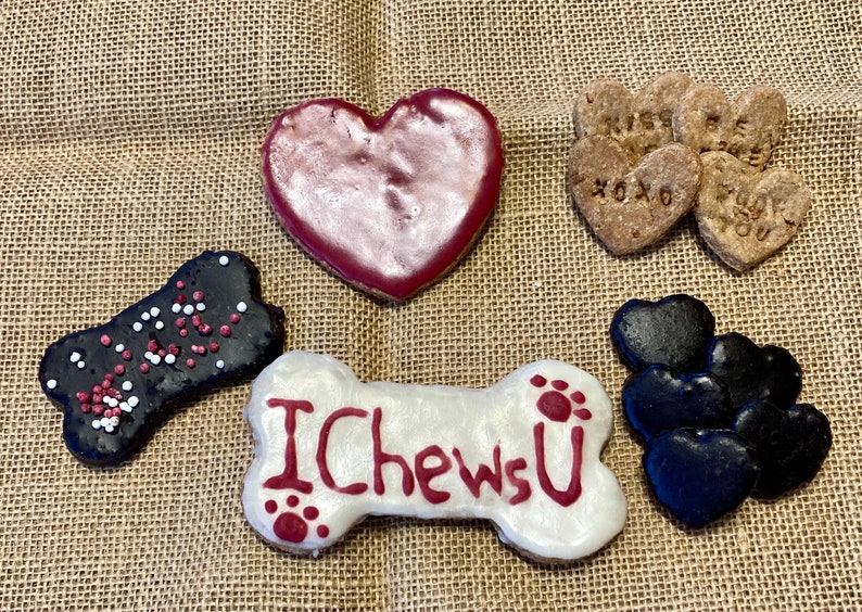 May include: Assortment of dog treats in the shape of hearts and bones. The treats are decorated with red, white and black icing and sprinkles. One bone-shaped treat has the words "I Chews U" written on it.