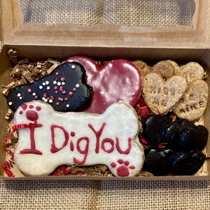 May include: A box of dog treats with a variety of shapes and flavours. The treats are decorated with red icing and sprinkles. The box includes a bone-shaped treat with the words "I Dig You" in red icing.