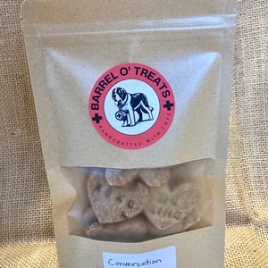 Pode incluir: Um saco de papel castanho com uma janela de pl&aacute;stico transparente que mostra guloseimas para c&atilde;es em forma de cora&ccedil;&atilde;o. O saco tem um logotipo vermelho e branco que diz "Barrel O' Treats + Handcrafted with Love". O saco tamb&eacute;m tem um r&oacute;tulo que diz "Conversation Hearts".
