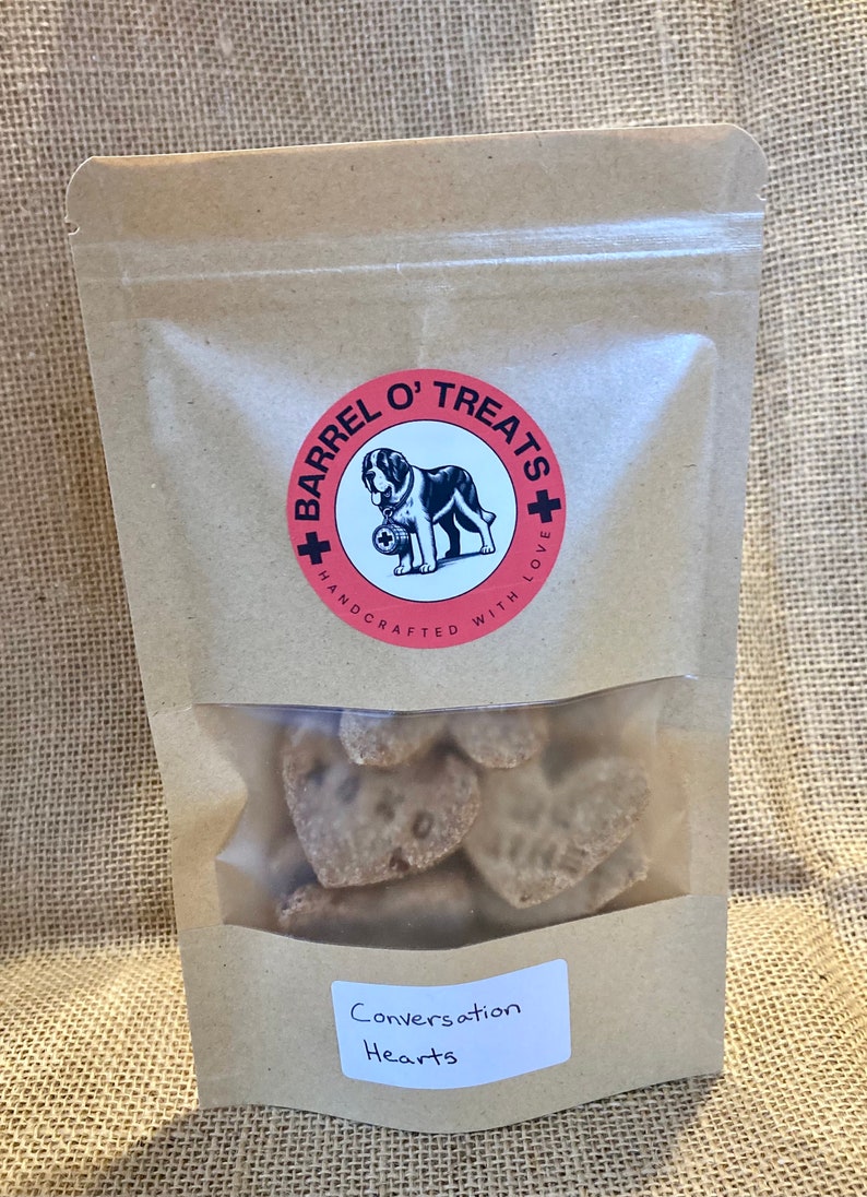 Pode incluir: Um saco de papel castanho com uma janela de pl&aacute;stico transparente que mostra guloseimas para c&atilde;es em forma de cora&ccedil;&atilde;o. O saco tem um logotipo vermelho e branco que diz "Barrel O' Treats + Handcrafted with Love". O saco tamb&eacute;m tem um r&oacute;tulo que diz "Conversation Hearts".