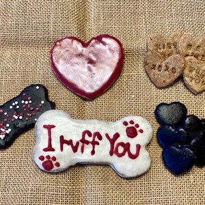 May include: Assortment of dog treats in the shape of hearts and bones. The treats are decorated with red, white and black icing and sprinkles. One bone-shaped treat has the words "I Ruff You" written on it.