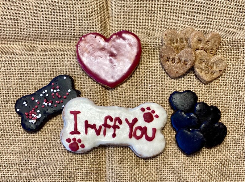 May include: Assortment of dog treats in the shape of hearts and bones. The treats are decorated with red, white and black icing and sprinkles. One bone-shaped treat has the words "I Ruff You" written on it.