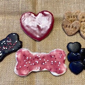 May include: Assortment of Valentine's Day dog treats. The treats are shaped like hearts and bones and are decorated with red and white icing and sprinkles. Some of the hearts have messages like "I miss you" and "XOXO".