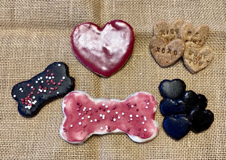 May include: Assortment of Valentine's Day dog treats. The treats are shaped like hearts and bones and are decorated with red and white icing and sprinkles. Some of the hearts have messages like "I miss you" and "XOXO".
