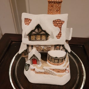 Può includere: Una casa di villaggio natalizio in ceramica bianca con una porta rossa, un tetto innevato e un camino in mattoni. La casa ha un design in stile pan di zenzero con una finestra e un cavallo a dondolo sul lato.