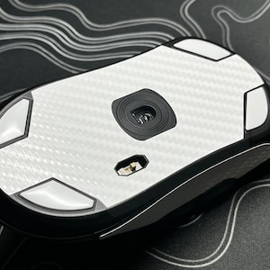 Könnte beinhalten: Weiße drahtlose Computermaus mit einem Karbonfaser-Muster auf Ober- und Unterseite. Die Maus hat vier weiße Füße auf der Unterseite und einen schwarzen Sensor in der Mitte.