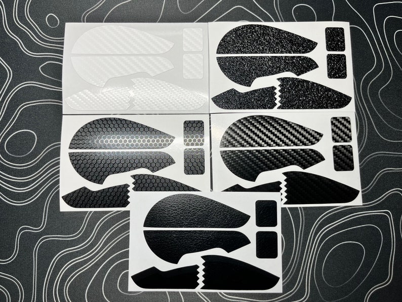 May include: A collection of mouse grip stickers in various designs. The stickers feature patterns like carbon fiber, textured black, and honeycomb. Each sticker set includes pieces for the mouse buttons and sides, all in black and white.