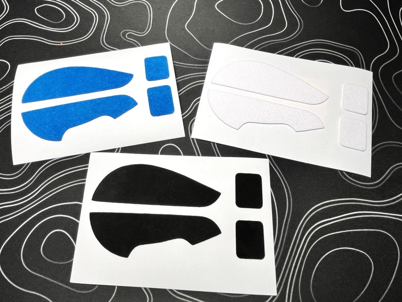 May include: Three sets of mouse grip stickers in blue, white, and black. Each set includes two larger pieces and two smaller square pieces, all on white backing. The blue stickers have a textured, glittery appearance.