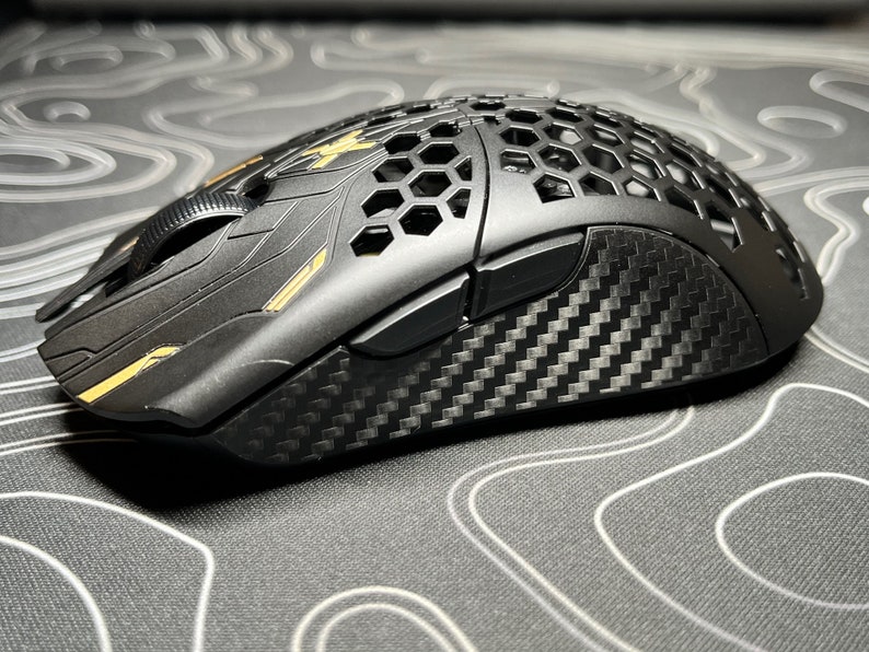 May include: Black gaming mouse with a honeycomb design on top. The mouse features carbon fiber accents and gold-colored details. The scroll wheel and side buttons are visible. The mouse is designed for gaming.