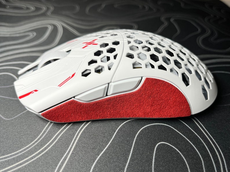 May include: White and red gaming mouse with a honeycomb design on the top. The mouse has red accents and a textured red grip on the side. The mouse has two side buttons and a scroll wheel.