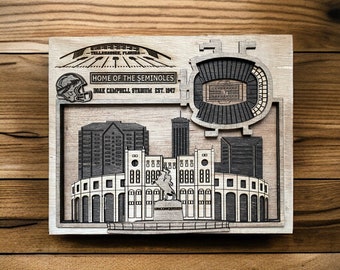 May include: A wooden wall art piece depicting a stylized cityscape of Tallahassee, Florida, with the text "HOME OF THE SEMINOLES DOAK CAMPBELL STADIUM EST. 1947". The artwork features a stadium, buildings, and a Seminole mascot.