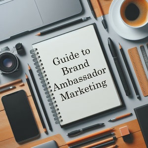 May include: A white notebook with a black spiral binding and the text "Guide to Brand Ambassador Marketing" written in black on the front. The notebook is surrounded by various office supplies, including pencils, pens, a ruler, a camera, and a smartphone.