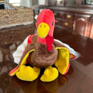 Puede incluir: Un juguete de peluche de pavo con cabeza roja, carúncula y plumas de la cola. El pavo tiene un cuerpo marrón, patas amarillas y alas amarillas y rojas. El juguete está sobre una superficie marrón.