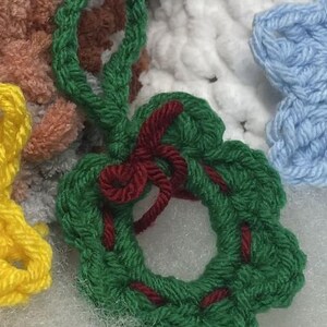 May include: A handmade green crochet wreath ornament with a red bow and accents. The wreath is round with a loop for hanging. Other colourful crochet items are visible in the background.