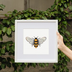 May include: A white frame with a white mat surrounding a cross-stitch of a yellow and black bee with clear wings.