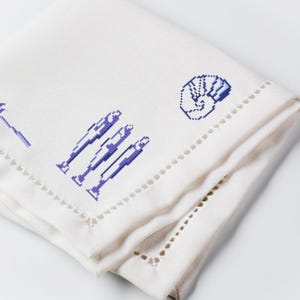 May include: White linen napkin with blue embroidered designs. The napkin features three stylized figures, a hand gesture, and a tool. The edges are finished with a decorative cut-out pattern.