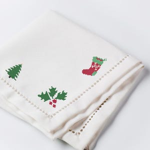 Puede incluir: Una servilleta de tela blanca doblada con diseños navideños bordados. Los diseños incluyen un árbol de Navidad verde, un calcetín rojo y verde y acebo verde con bayas rojas. Los bordes tienen un patrón decorativo recortado.