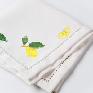 Puede incluir: Servilleta de lino blanco con diseño de limón y hojas bordado a punto de cruz. El limón es amarillo con hojas verdes, y la servilleta tiene un borde decorativo calado. La servilleta está doblada, mostrando el diseño en la capa superior.