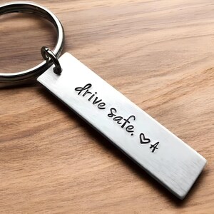 Puede incluir: Llavero de plata con una etiqueta rectangular que dice "drive safe. ♥ A".