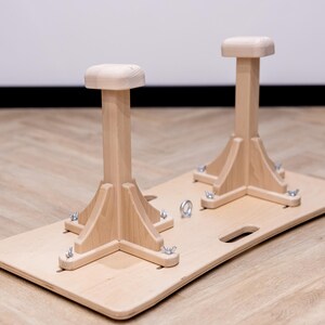 May include: A wooden gymnastics training aid featuring two upright posts with square tops, mounted on a rectangular base. The natural wood color is accented by metal hardware. The base has a handle cutout.
