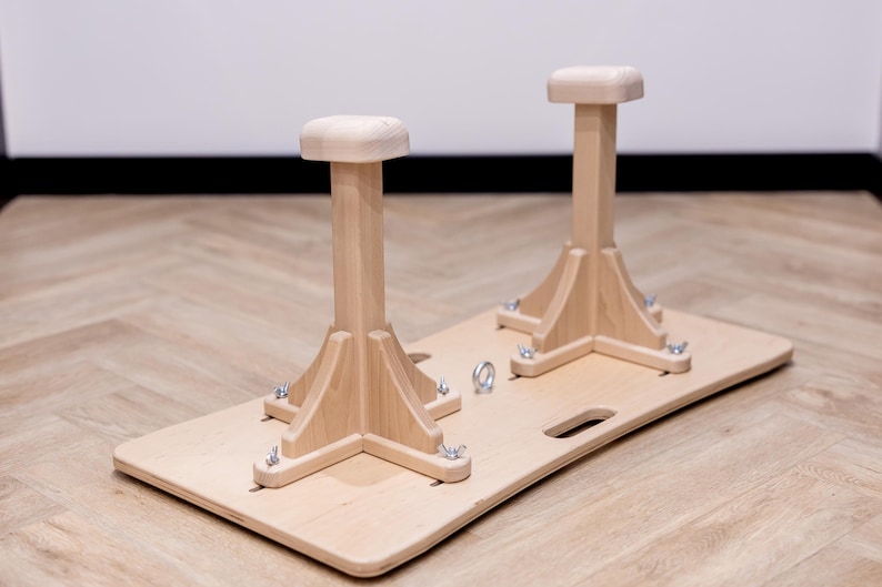 May include: A wooden gymnastics training aid featuring two upright posts with square tops, mounted on a rectangular base. The natural wood color is accented by metal hardware. The base has a handle cutout.