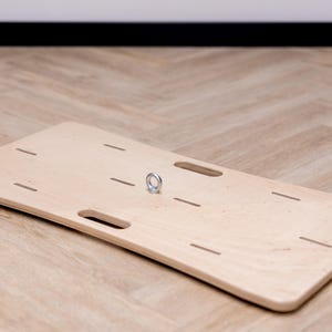 May include: A light-colored wooden balance board with a metal ring and several cut-out slots. The board is rectangular with rounded corners and two handle cutouts. The board is on a light brown wooden floor.