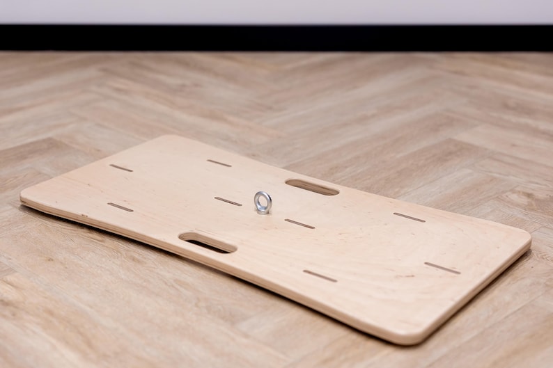 May include: A light-colored wooden balance board with a metal ring and several cut-out slots. The board is rectangular with rounded corners and two handle cutouts. The board is on a light brown wooden floor.