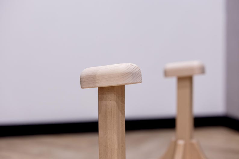 May include: Two wooden handstand blocks with rounded tops and sturdy, vertical supports. The blocks are made of light-colored wood and are designed for balance and stability during handstand practice. The background is a plain white wall.