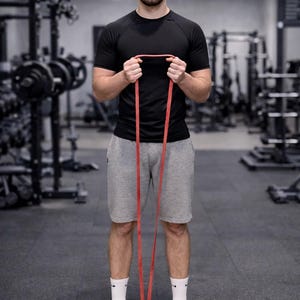 May include: A person in a gym setting holding a red resistance band. The person is wearing a black t-shirt, gray shorts, and white socks. They are standing on a wooden balance board.
