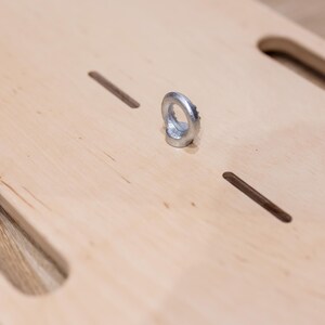 May include: A light-colored wooden board with two large, rounded rectangular cutouts and two smaller, rectangular slots. A silver metal ring is attached to the board. The board's surface has a natural wood grain.