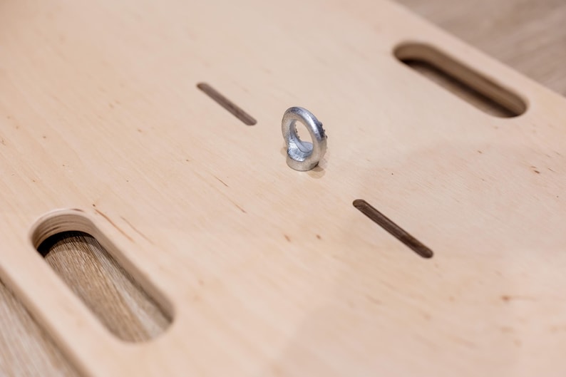 May include: A light-colored wooden board with two large, rounded rectangular cutouts and two smaller, rectangular slots. A silver metal ring is attached to the board. The board's surface has a natural wood grain.