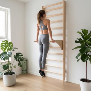 Könnte beinhalten: Eine hölzerne Kletterleiter zur Wandmontage mit horizontalen Sprossen. Die Leiter ist hell und an einer weißen Wand angebracht. Eine Person trainiert mit der Leiter, bekleidet mit einem grauen Sport-BH und Leggings. Der Raum hat Pflanzen und einen Holzboden.