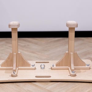 May include: A wooden gymnastics training aid. The apparatus features two vertical posts with rounded tops, mounted on a rectangular base. The natural wood color and simple design suggest a focus on functionality and natural materials.