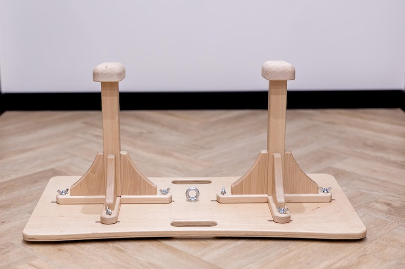 May include: A wooden gymnastics training aid. The apparatus features two vertical posts with rounded tops, mounted on a rectangular base. The natural wood color and simple design suggest a focus on functionality and natural materials.