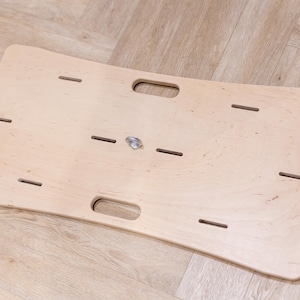 May include: A light-colored wooden balance board with a curved design. It features two handle cutouts and several rectangular slots. A metal swivel is in the center. The board is designed for exercise and physical therapy.