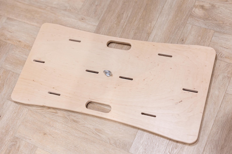 May include: A light-colored wooden balance board with a curved design. It features two handle cutouts and several rectangular slots. A metal swivel is in the center. The board is designed for exercise and physical therapy.