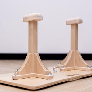 May include: Wooden gymnastics handstand blocks. Two light-colored wooden handstand blocks are attached to a rectangular wooden base. The blocks have a rectangular top and a flared base. The base has metal hardware.