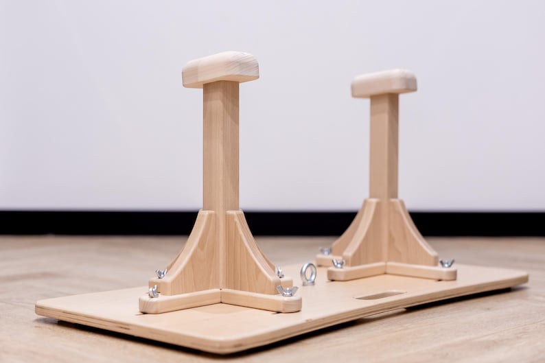 May include: Wooden gymnastics handstand blocks. Two light-colored wooden handstand blocks are attached to a rectangular wooden base. The blocks have a rectangular top and a flared base. The base has metal hardware.