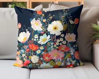 Puede incluir: Un cojín decorativo cuadrado con un diseño floral. El cojín presenta un fondo azul oscuro con una variedad de flores en tonos blancos, rosas, rojos y amarillos. El cojín está colocado sobre un sofá gris.