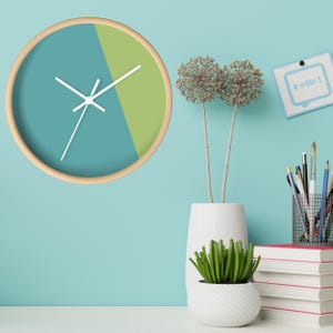 May include: A round wall clock with a light wood frame. The clock face is divided into two sections: teal and lime green. White hands indicate the time. The background is a light blue wall. A small plant and a stack of books are on a white surface.