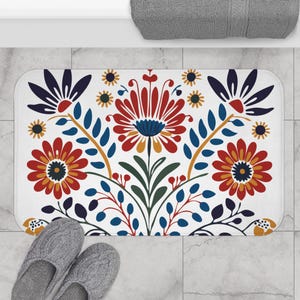 May include: A white bath mat with a colorful floral design. The flowers are red, yellow, blue, and green. The design is symmetrical and has a traditional folk art style.