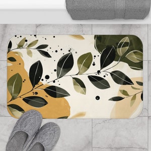 Puede incluir: Una alfombra de baño blanca con un diseño de hojas en tonos verdes, amarillos y marrones. Las hojas son estilizadas y abstractas, con un efecto acuarela. La alfombra tiene un respaldo antideslizante.