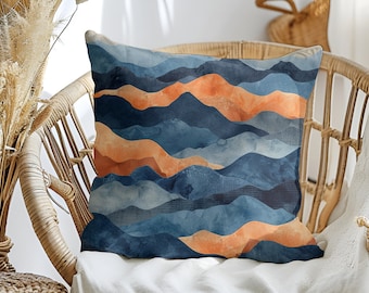 Könnte beinhalten: Ein dekoratives Kissen mit einem abstrakten Bergdesign in Blau und Orange. Das Kissen liegt auf einem Korbstuhl.