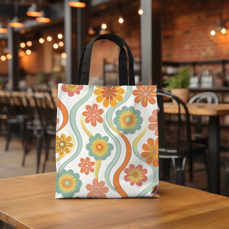 May include: A white tote bag with a colorful floral pattern. The design features orange, yellow, and pink flowers with green and orange wavy lines. The bag has black handles and is sitting on a wooden table.