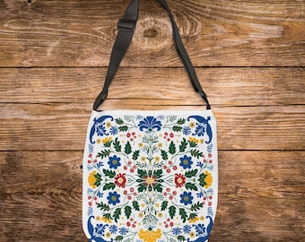 May include: White crossbody bag with a floral pattern in blue, red, yellow, and green. The bag has a black adjustable strap and a square shape. The floral design is symmetrical and detailed.