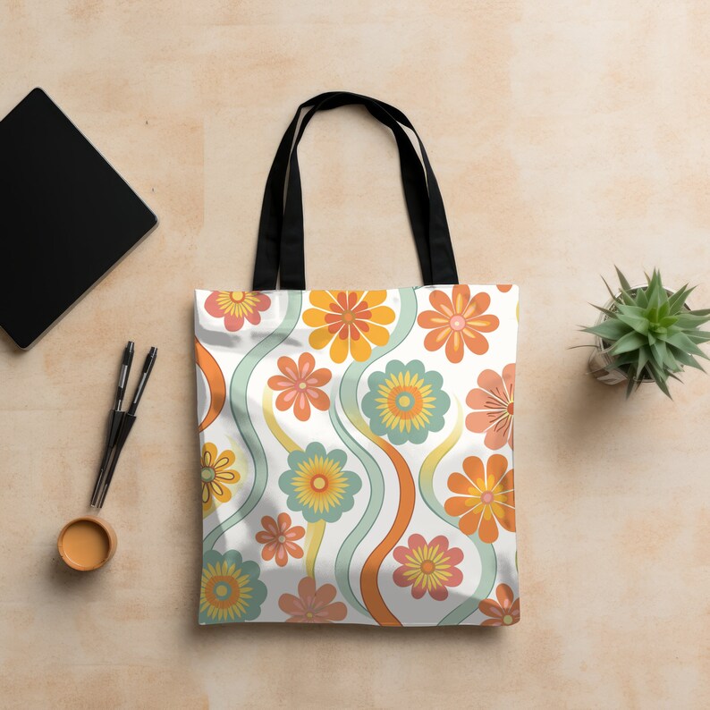 May include: White tote bag with a retro floral pattern in orange, yellow, and teal. The bag has black handles and a simple design. The bag is sitting on a light brown surface.