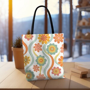 May include: A white tote bag with a colorful floral pattern. The design features orange, yellow, and pink flowers with green and orange wavy lines. The bag has black handles and is sitting on a wooden surface.