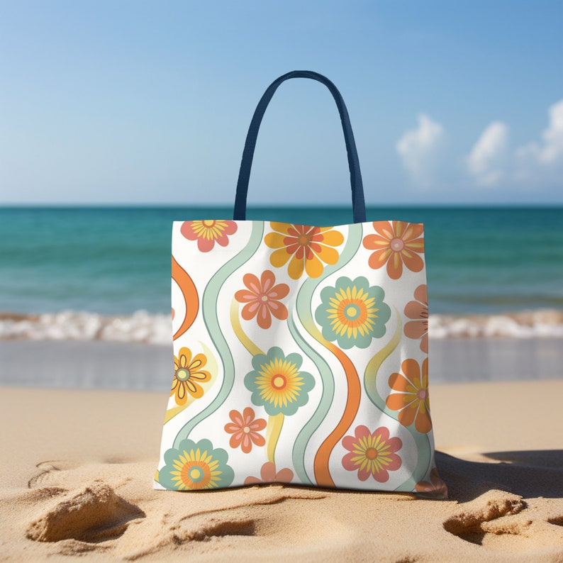 May include: A tote bag with a retro floral pattern in orange, yellow, and teal on a white background. The bag has a navy blue handle and is sitting on a sandy beach with the ocean in the background.